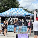A group of people are gathered around a food stand, waiting to be served authentic German sausage.