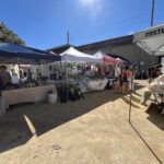 A group of people are gathered around a tent, possibly at a market or event.