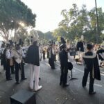 A group of people in uniforms are standing on a street corner, possibly a marching band, as they prepare to perform.