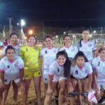 A group of women pose together on a beach, wearing matching uniforms and holding their hands together.