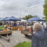 A large group of people are gathered around a market, with a variety of items on display.