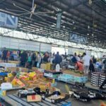 A large group of people are gathered in a warehouse, browsing through a variety of items, including boxes of apples and oranges.