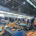 A large group of people are shopping in a market, with various items such as apples, oranges, and bottles.