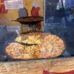 A large pan of food is displayed in a window, with a sign that says "Just say my name."