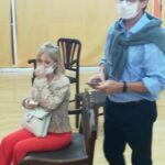 A man and woman are standing in a room, with the woman sitting on a chair and the man standing next to her. The woman is wearing a face mask, and the man is holding a cell phone.