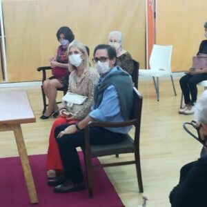 A man and woman wearing face masks sit in chairs, possibly in a classroom setting.