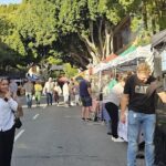 A man in a black shirt stands in front of a food stand, smiling as he interacts with the woman beside him.