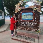 A man in a red jacket stands in front of a sign for Ushuaia, the southernmost city in the world.