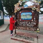 A man in a red jacket stands in front of a sign for Ushuaia, the southernmost city in the world.