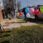 A man is hanging balloons on a pole, while a teddy bear sits nearby.
