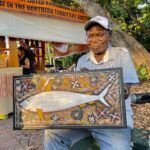 A man is sitting on a bench holding a painting of a fish.
