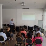 A man stands in front of a projector screen, presenting to a group of people.