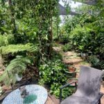 A patio table with a blue tablecloth and a chair sits in a garden with a variety of plants and flowers.