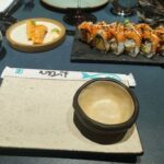 A table with a variety of foods, including sushi and sashimi, and a bowl with a spoon in it.