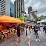 A woman in a black dress walks past a fruit juice stand at a market.