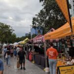 A woman in an orange shirt stands in front of a food stand at a market.