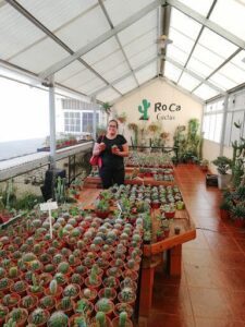 A woman is standing in a room filled with potted plants, holding a small plant in her hand.
