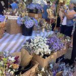 A woman is standing in front of a table filled with flowers, including purple and blue ones.