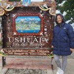 A woman stands in front of a sign that reads "Ushuaia fin del mundo."