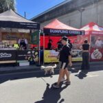 A woman walks her dog on a leash while passing by a food stand.