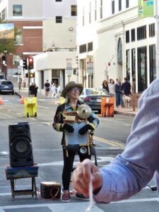A woman wearing a hat and glasses is standing on a street corner holding a drum.