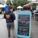 A young boy wearing a jacket with a strawberry on it stands in front of a chalkboard sign advertising a strawberry shortcake.
