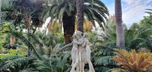 Three statues of people are standing together in a garden, surrounded by palm trees.