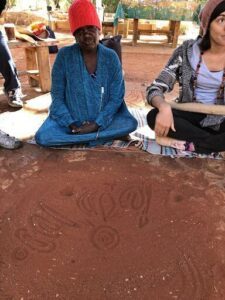 Two women sit on the ground, one of them is drawing a picture in the dirt.