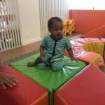 A baby is sitting on a green and red block, possibly in a play area.
