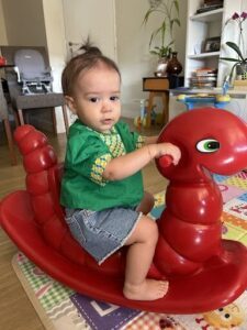 A baby is sitting on a red snake toy, wearing a green shirt.