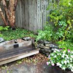 A bench sits in front of a stone wall, surrounded by flowers and plants.
