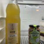 A bottle of corumma sits next to a jar of kimchi on a shelf.
