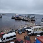 A busy harbor with boats and cars, including a red car, a white van, and a truck.