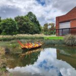 A colorful boat made of glass sits in a pond, reflecting the sky above.