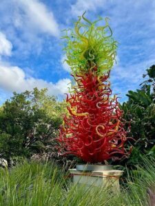 A colorful, tall glass sculpture stands in a field, surrounded by greenery.