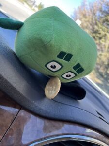 A green stuffed animal is sitting on a car's dashboard, with a cracker in its mouth.