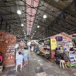 A group of people are walking through a market, with a man sitting on a chair in the foreground.