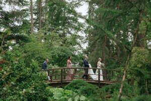 A group of people, including a bride and groom, walk across a bridge in a forest.