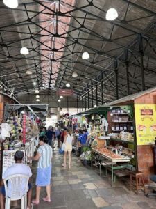 A group of people walking through a market, with a woman in a striped shirt leading the way.