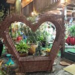 A heart-shaped woven basket sits on a patio, surrounded by potted plants and a bench.