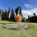 A large, colorful sculpture of a tree is displayed in a park, surrounded by a variety of plants and flowers.