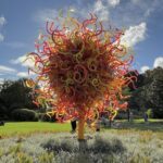 A large, colorful sculpture of a tree stands in a park, surrounded by people enjoying the day.