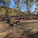 A large group of people are gathered in a field, sitting on the ground and enjoying a day outdoors.