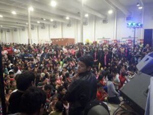 A large group of people are gathered in a room, with some standing and others sitting. The crowd is diverse, and they appear to be enjoying a social event.