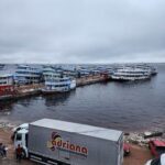 A large truck is parked near a body of water, with several boats docked nearby.