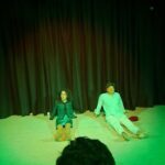 A man and a woman are lying on the floor, possibly in a theatrical setting.