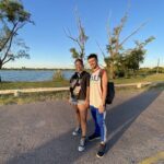 A man and woman pose for a picture on a road near a body of water.