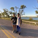 A man and woman stand on a road near a lake, posing for a picture.