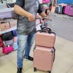A man in a black shirt is standing next to a pink suitcase, giving a thumbs up.
