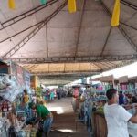 A man in a white shirt is shopping at a market, surrounded by various items such as bottles, a banana, and a bowl.